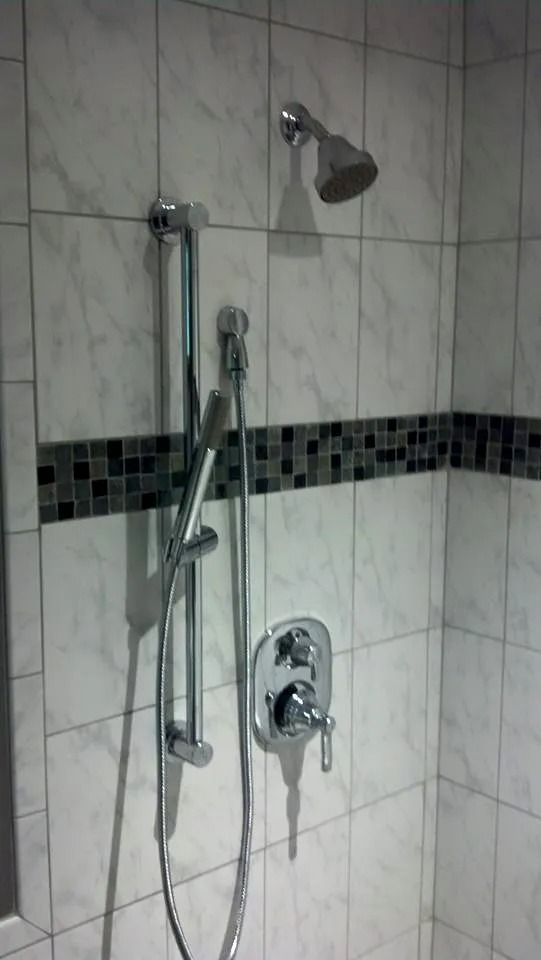 A tiled shower wall with a chrome fixed showerhead, a sliding handheld sprayer, and a single-lever mixing valve.