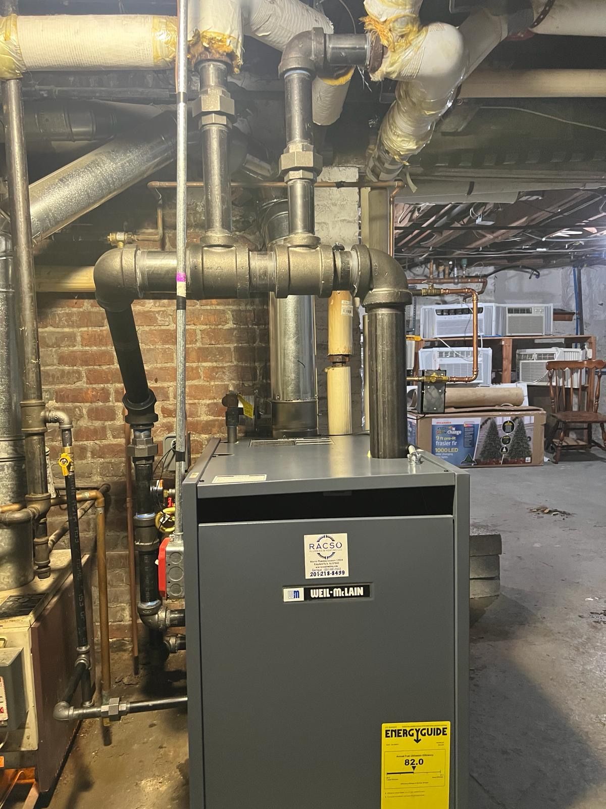 A boiler is sitting in a basement next to pipes and a brick wall