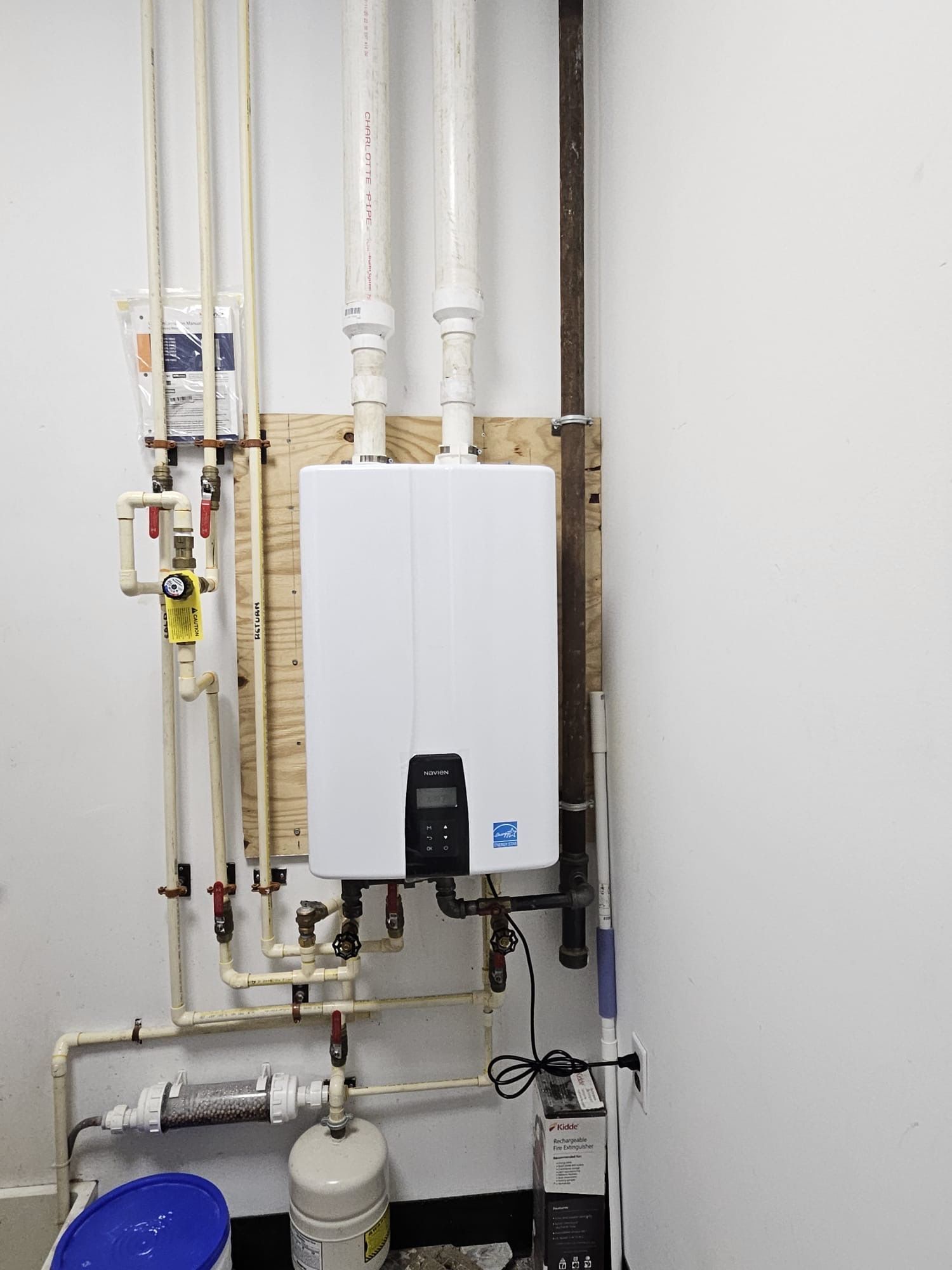 A white boiler is mounted to the wall in a room with pipes