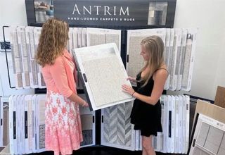 Two women looking at carpet samples in a showroom. They are examining a large, neutral-colored carpet square.