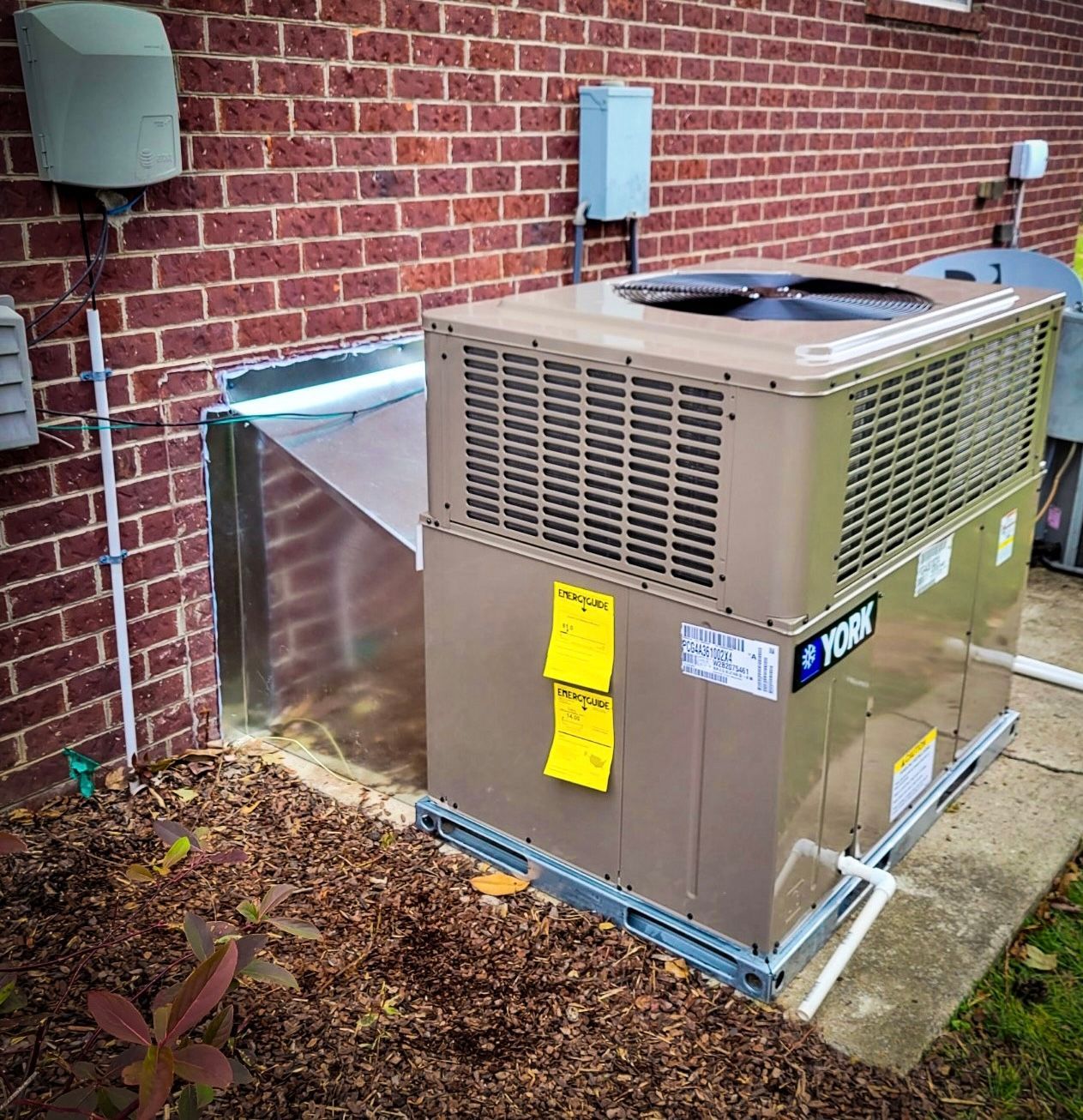 Exterior HVAC unit beside a brick building with ductwork and utility boxes.