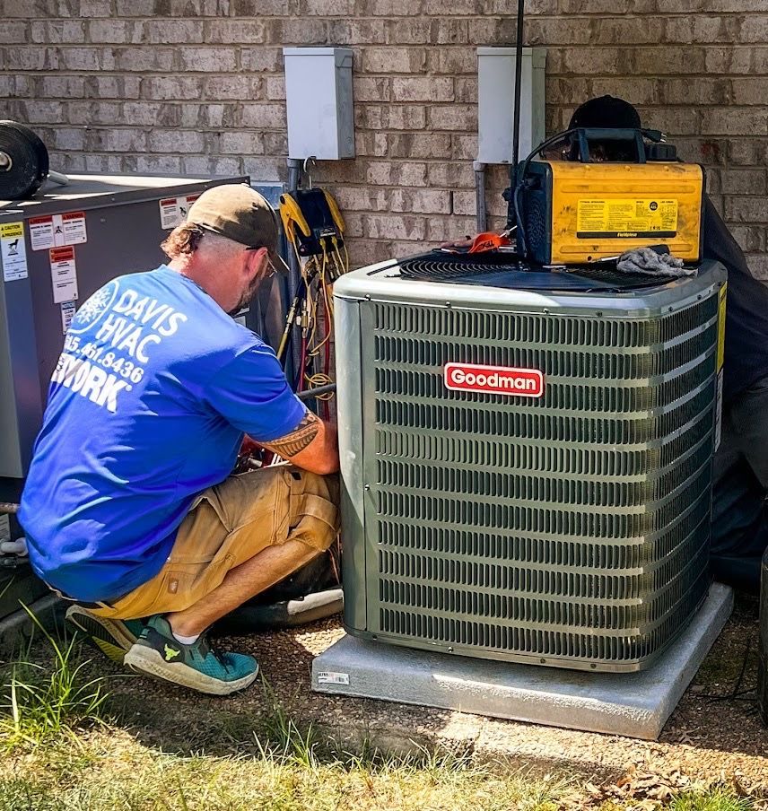 HVAC technician working on an outdoor Goodman air conditioning unit. Brick wall background.