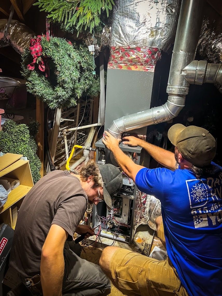 Two technicians working on HVAC system in an attic; one in blue shirt, one in brown.