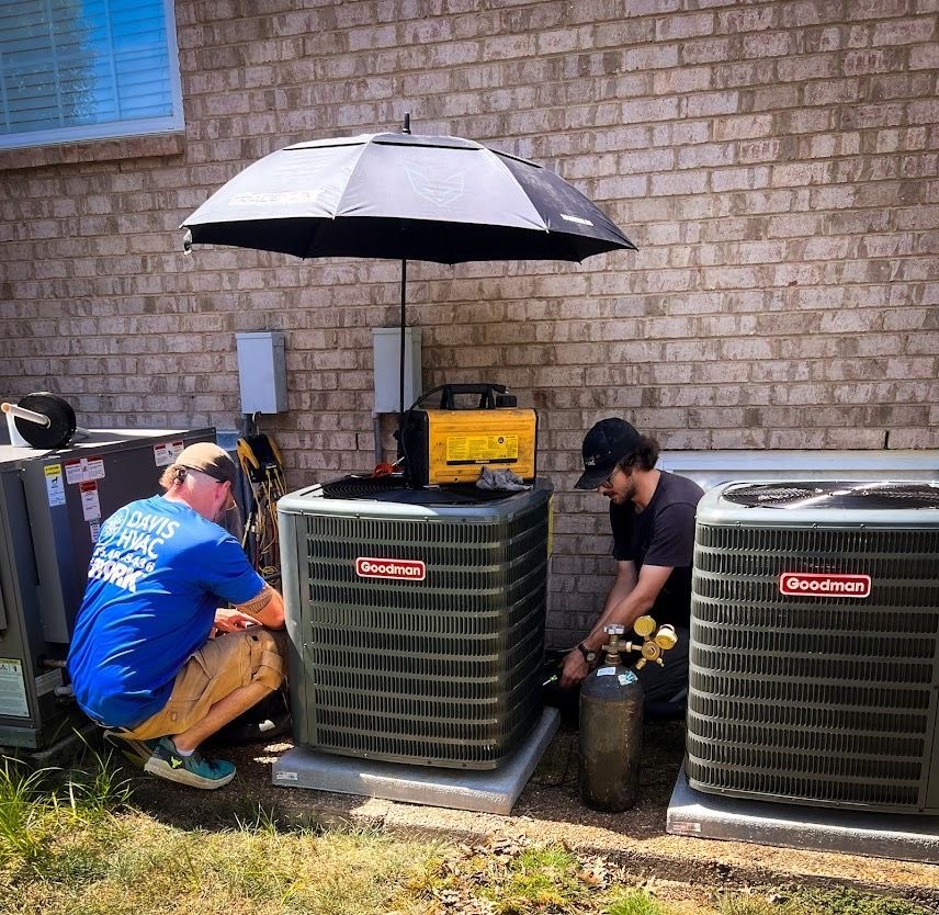 Two HVAC technicians working on an air conditioning unit outside, under an umbrella.