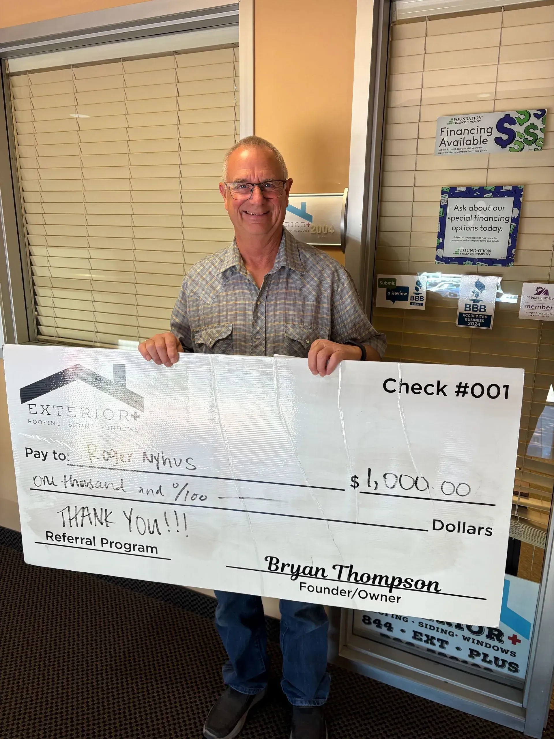 Man holding an oversized check indoors, standing by a window and bulletin board