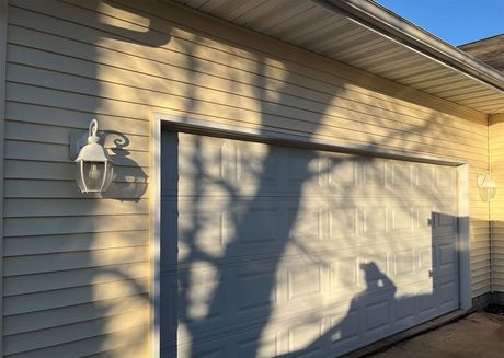 Closed garage door on a beige house exterior with a wall-mounted light fixture and tree shadows.