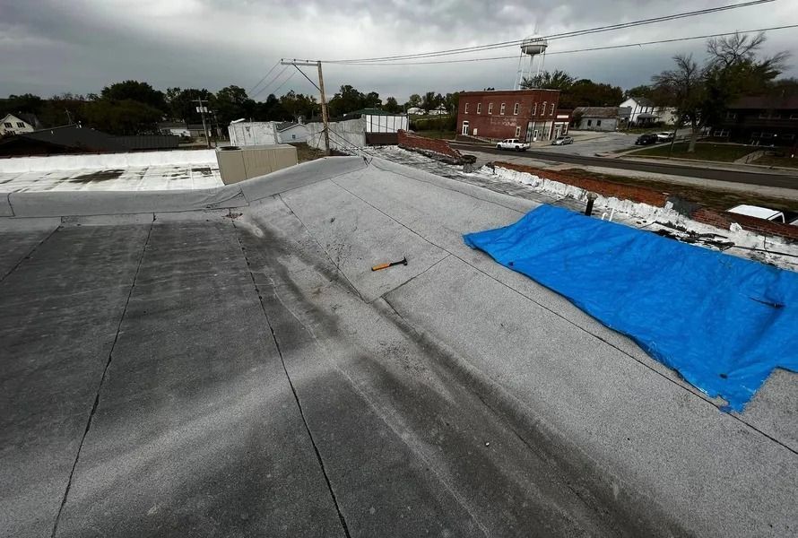 Flat rooftop under repair with gray membrane and a blue tarp beside a brick building in the distance