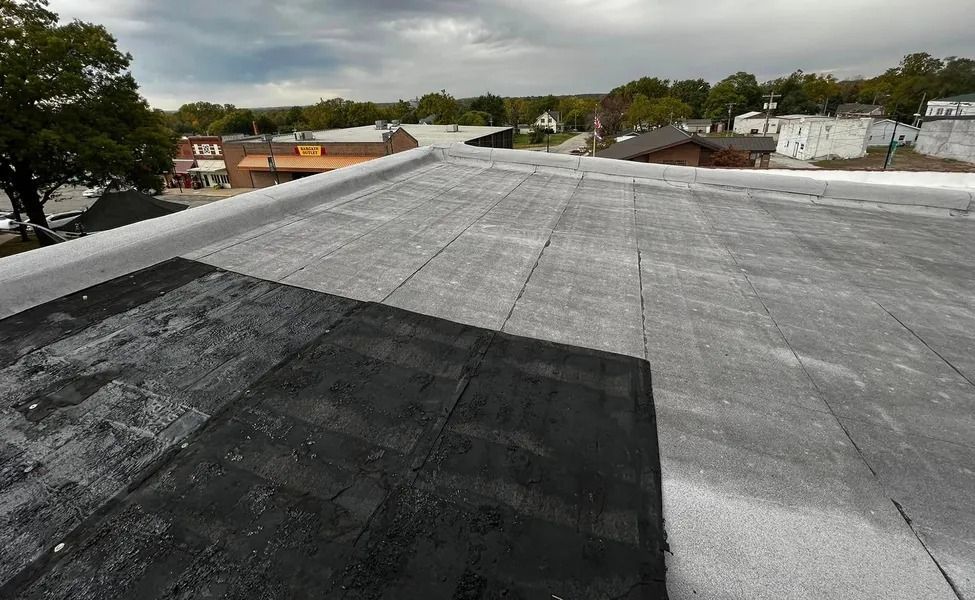 Flat rooftop with gray and black roofing under cloudy skies, overlooking nearby trees and buildings.