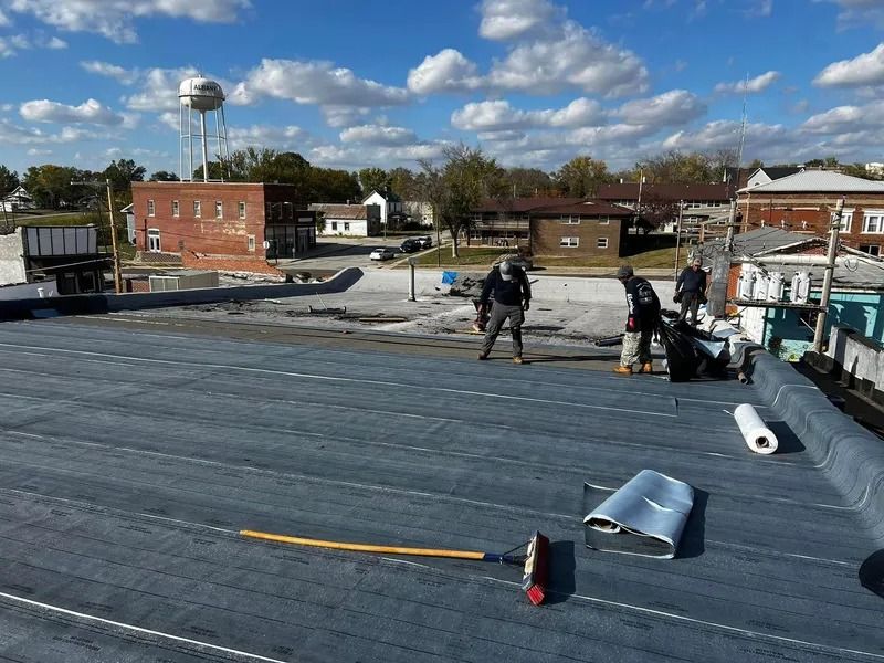 Workers on a flat rooftop installing materials beside rolled roofing under a partly cloudy sky