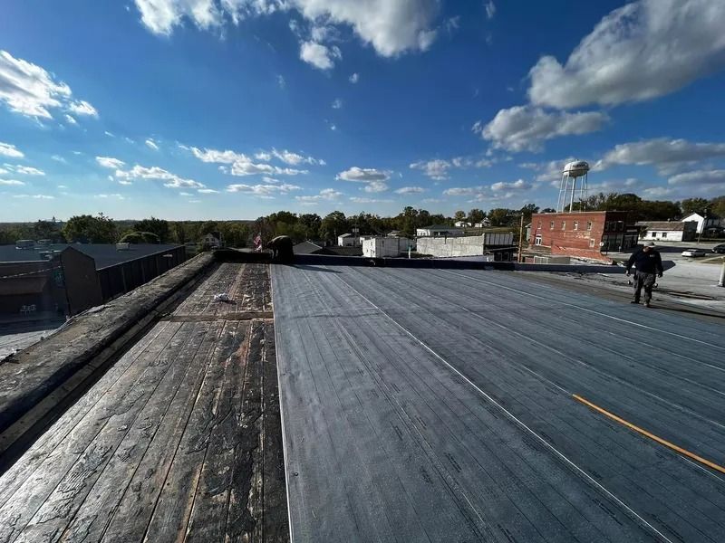 Rooftop workers installing a dark membrane on a flat roof under a bright blue sky.