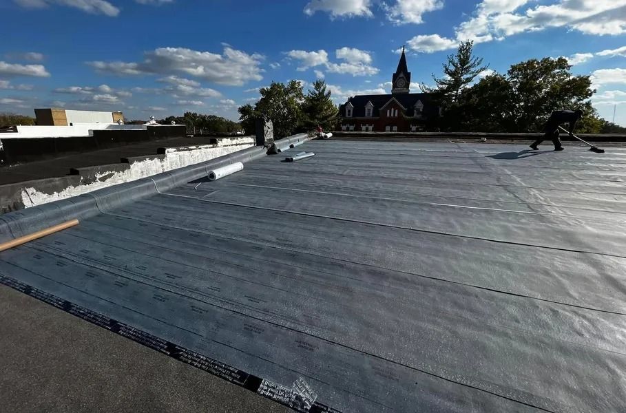 Flat rooftop covered in dark membrane under a blue sky, with buildings and a church steeple in the distance.
