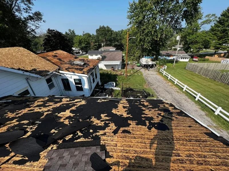 Storm-damaged roof with missing shingles on a white house in a sunny suburban neighborhood.