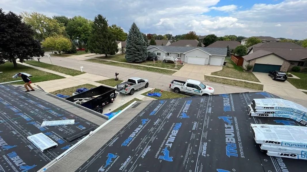 Roofer installing black underlayment on a house roof in a suburban neighborhood under a cloudy sky