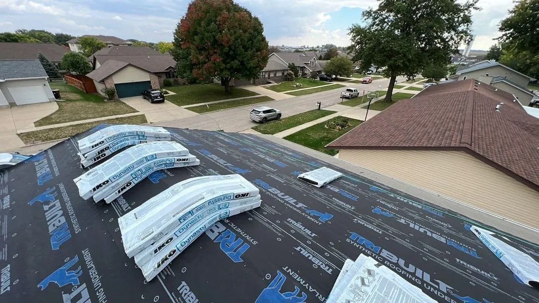 Roof under repair with underlayment and stacked shingles, overlooking a suburban street and houses.