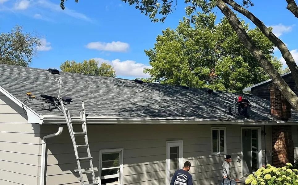 Workers repairing a house roof, with ladders and shingles under a blue sky.