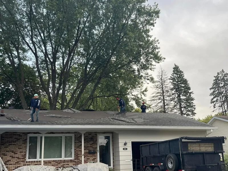 Workers on a house roof clearing debris beside a black dumpster, with trees in the background.