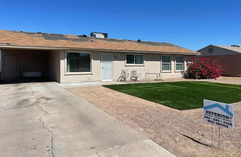 Single-story beige house with carport, driveway, green lawn, and a real estate sign in front.