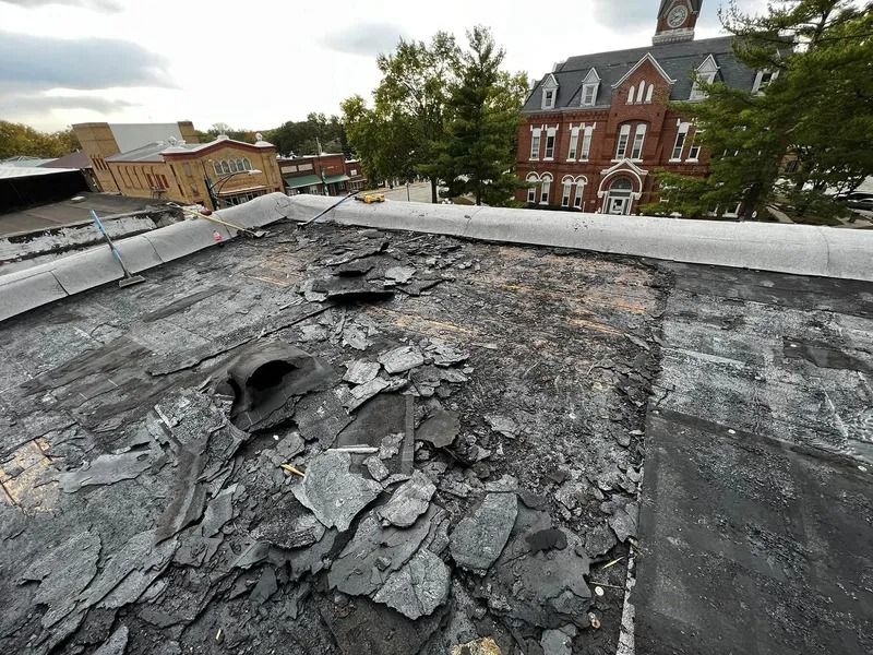 Damaged flat roof with peeling tar and debris, overlooking buildings and trees in the background