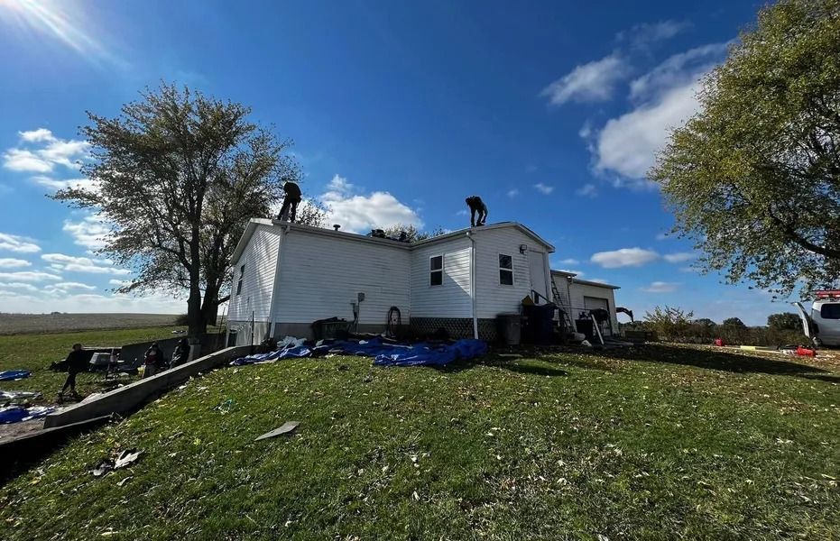 White house with damaged roof on a grassy hill under a blue sky, with trees and scattered debris nearby