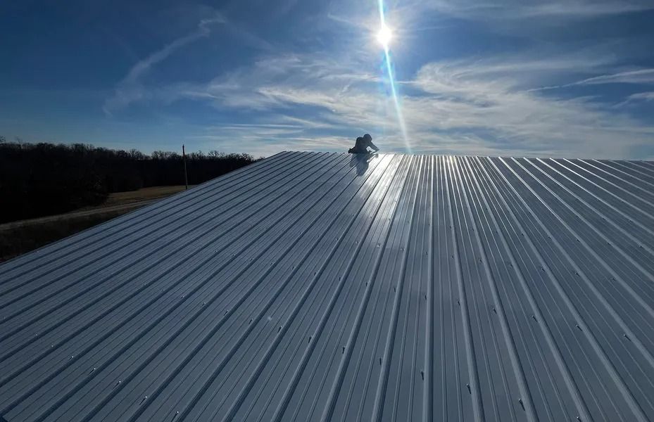 Worker on a sunlit corrugated metal roof under a blue sky, with trees in the distance.