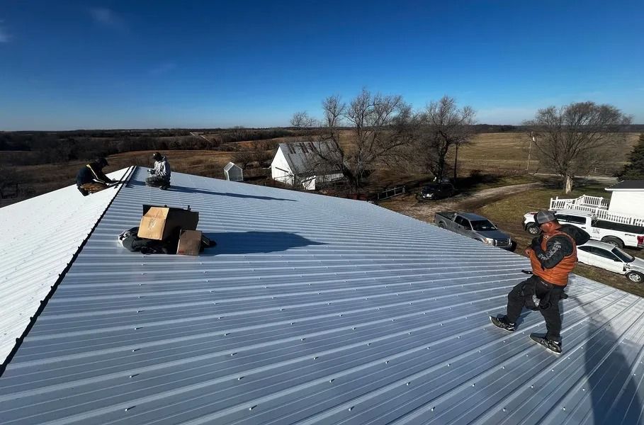 Worker on a metal roof using a power tool, with roofing materials and rural landscape in the background.