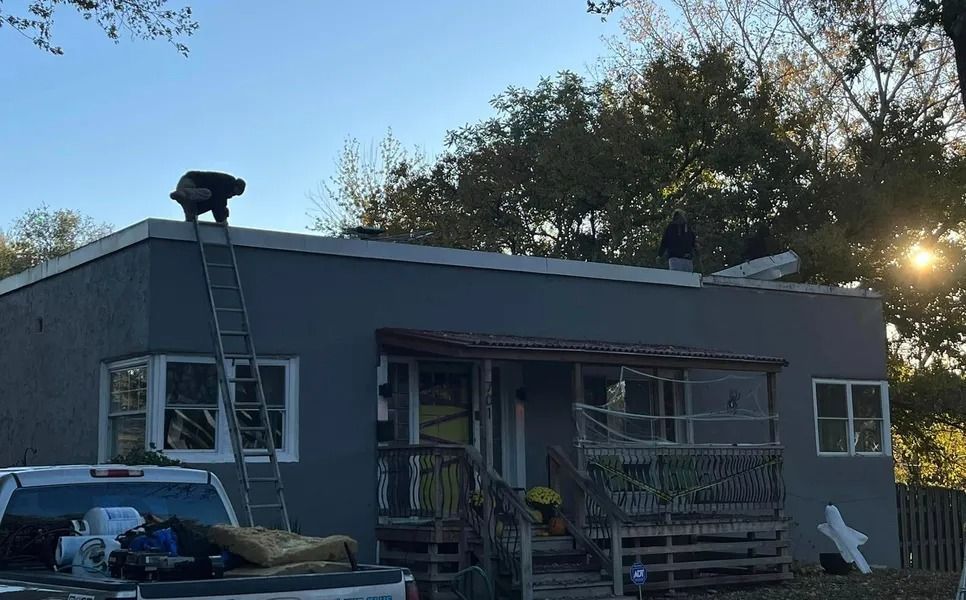 Dark gray house with damaged roof and cluttered yard at dusk, sun setting behind trees.
