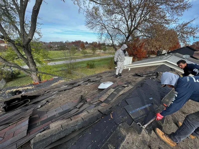 Workers repair a damaged shingle roof beside trees and a rural road under a cloudy sky