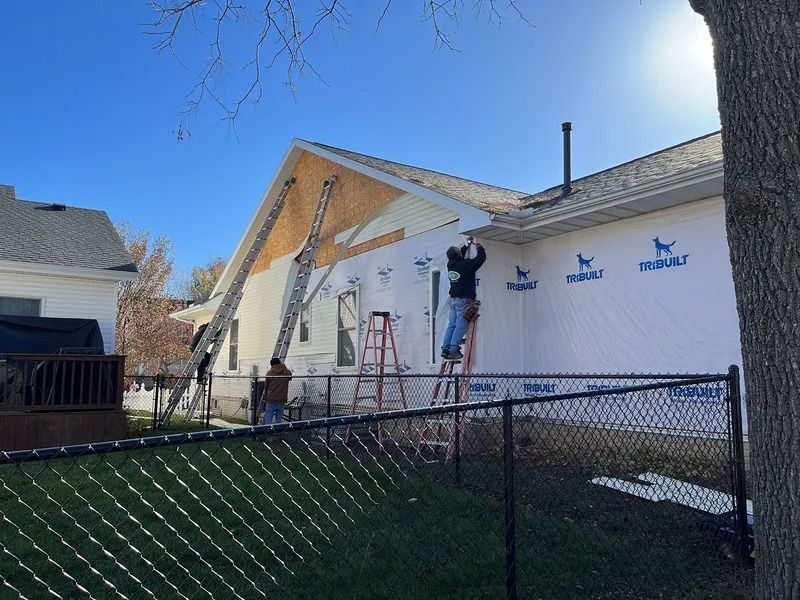 Workers installing house wrap on a white house beside a chain-link fence on a sunny day