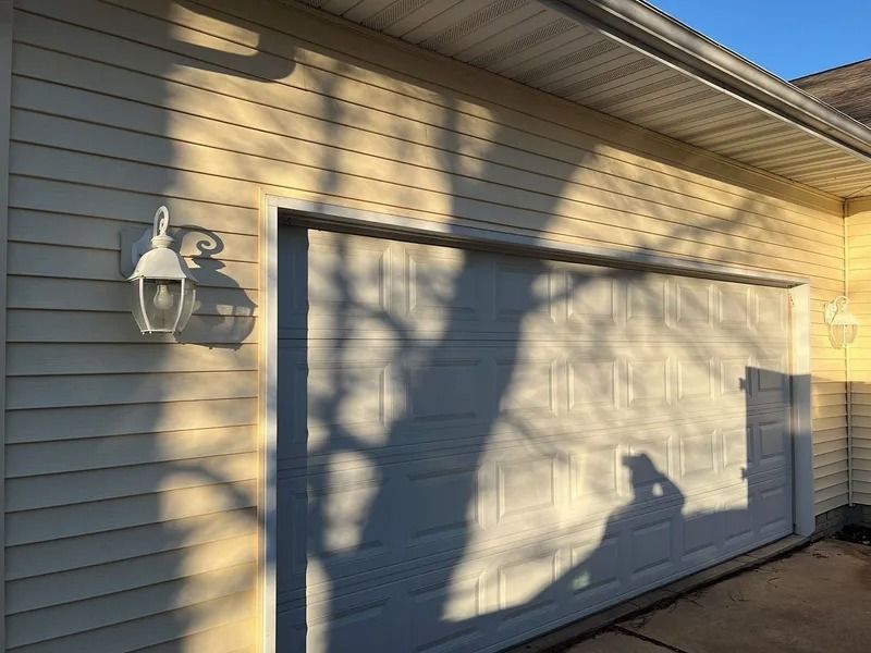 Garage door with tree shadows on beige siding beside an exterior wall light.