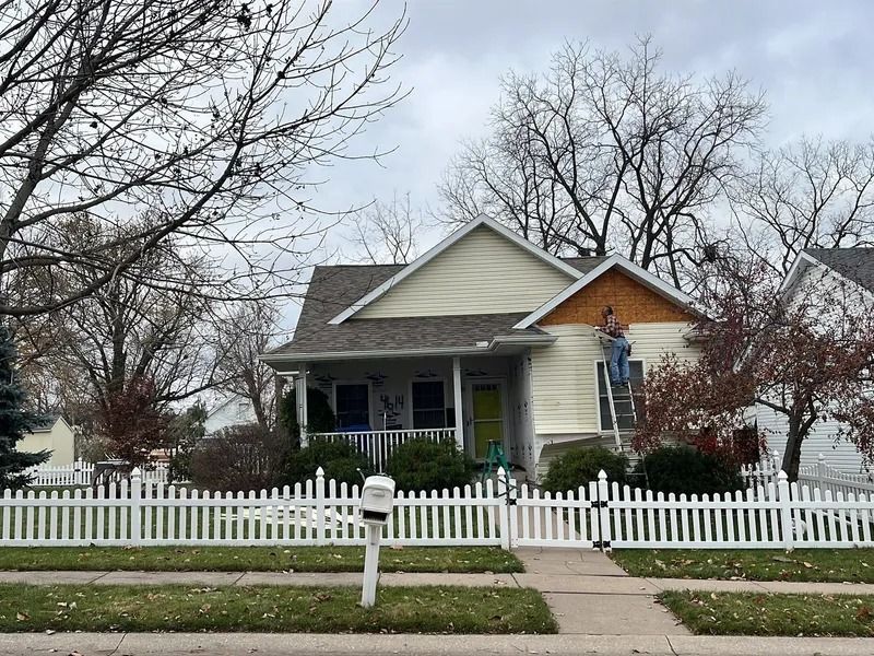 House with white picket fence, front porch, and ladder by the roof in a leafless yard.