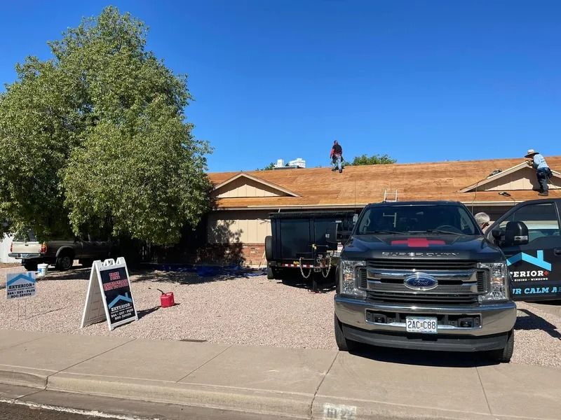Front yard with two vehicles, a roof being repaired, and a large tree under a clear blue sky.