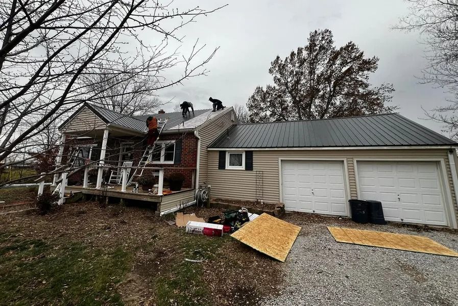 Damaged house with roof work in progress, debris on the yard, and two people on the roof.