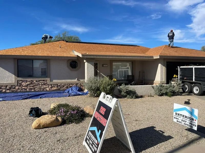 Single-story house with orange roof, gravel yard, and yard signs under a blue sky; worker on the roof.