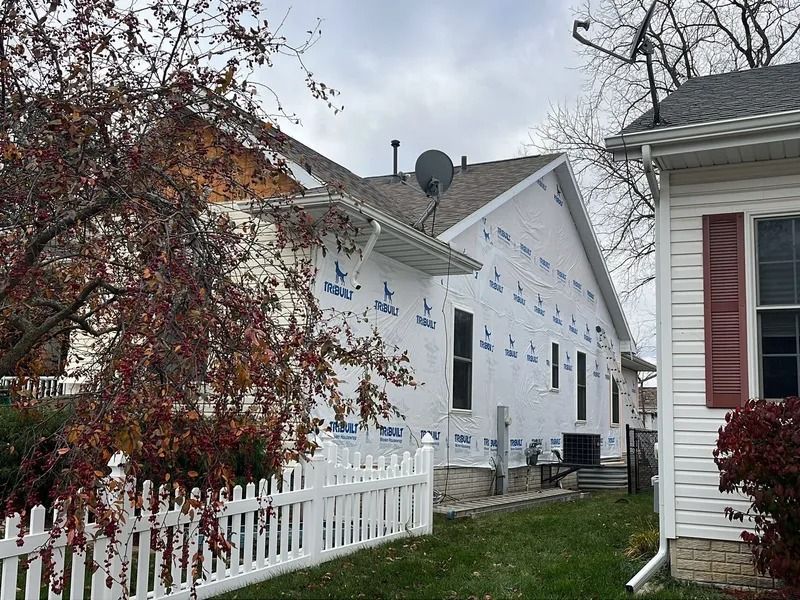 White house under construction with house wrap, white picket fence, and leafless trees in a residential yard