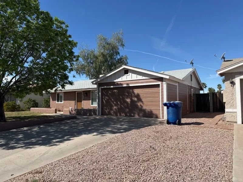 Suburban driveway with a tan garage, blue recycling bin, gravel yard, and trees under a clear sky
