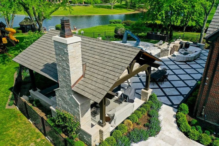 Aerial view of a stone house with a gray roof beside a patio, lawn, and pond in a landscaped yard