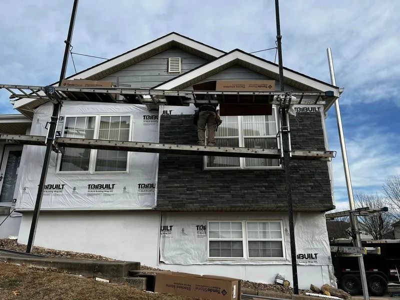 Damaged two-story house under repair, with exposed framing, scaffolding, and wrapped siding.