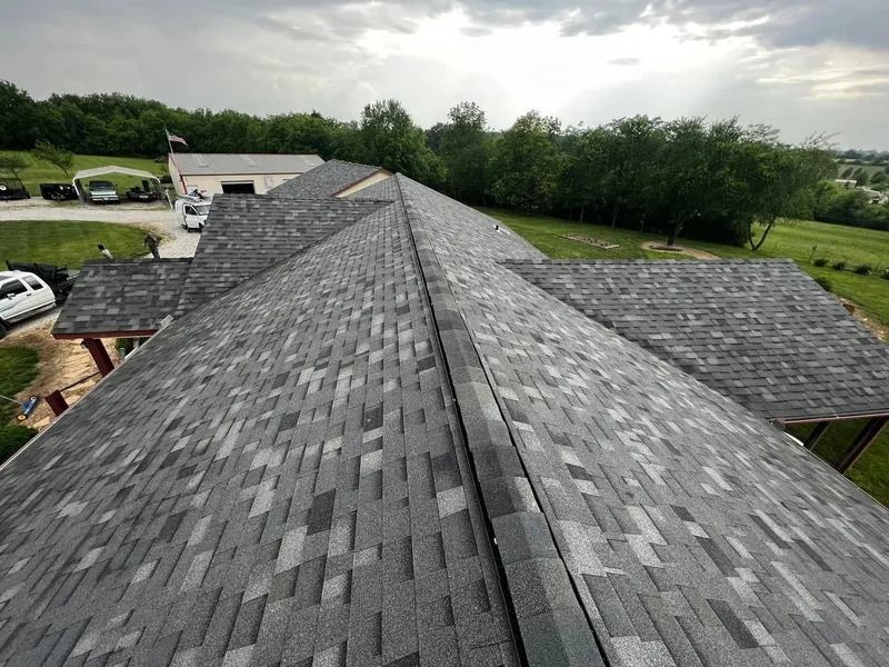 Aerial view of a gray shingled roof on a house surrounded by green trees and cloudy sky.