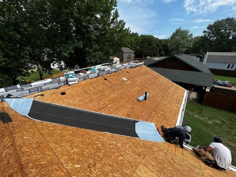 Workers installing shingles on a large orange roof, with trees and neighboring houses nearby