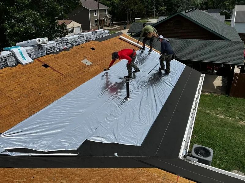Workers installing a silver roof underlayment on a house roof, with shingles stacked nearby.