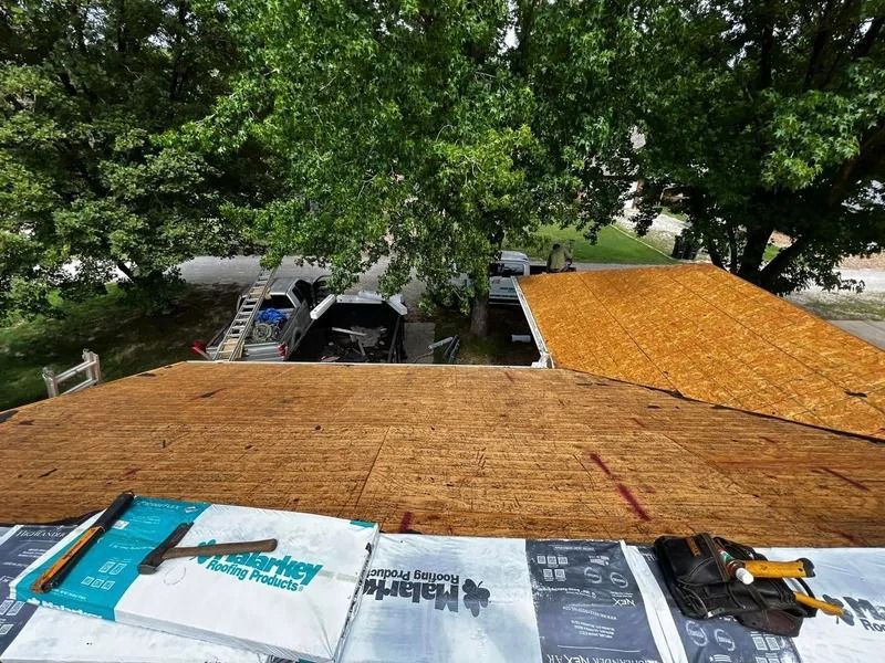 Roof under repair with exposed plywood and roofing materials, viewed from above amid trees