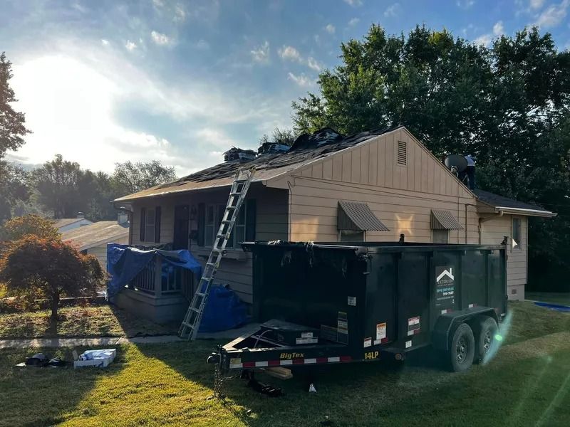 House roof under repair with ladder, debris trailer, and dumpster in a sunny yard