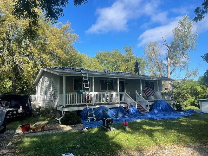 A house under repair with ladders, blue tarps, and a person on the roof in a sunny yard