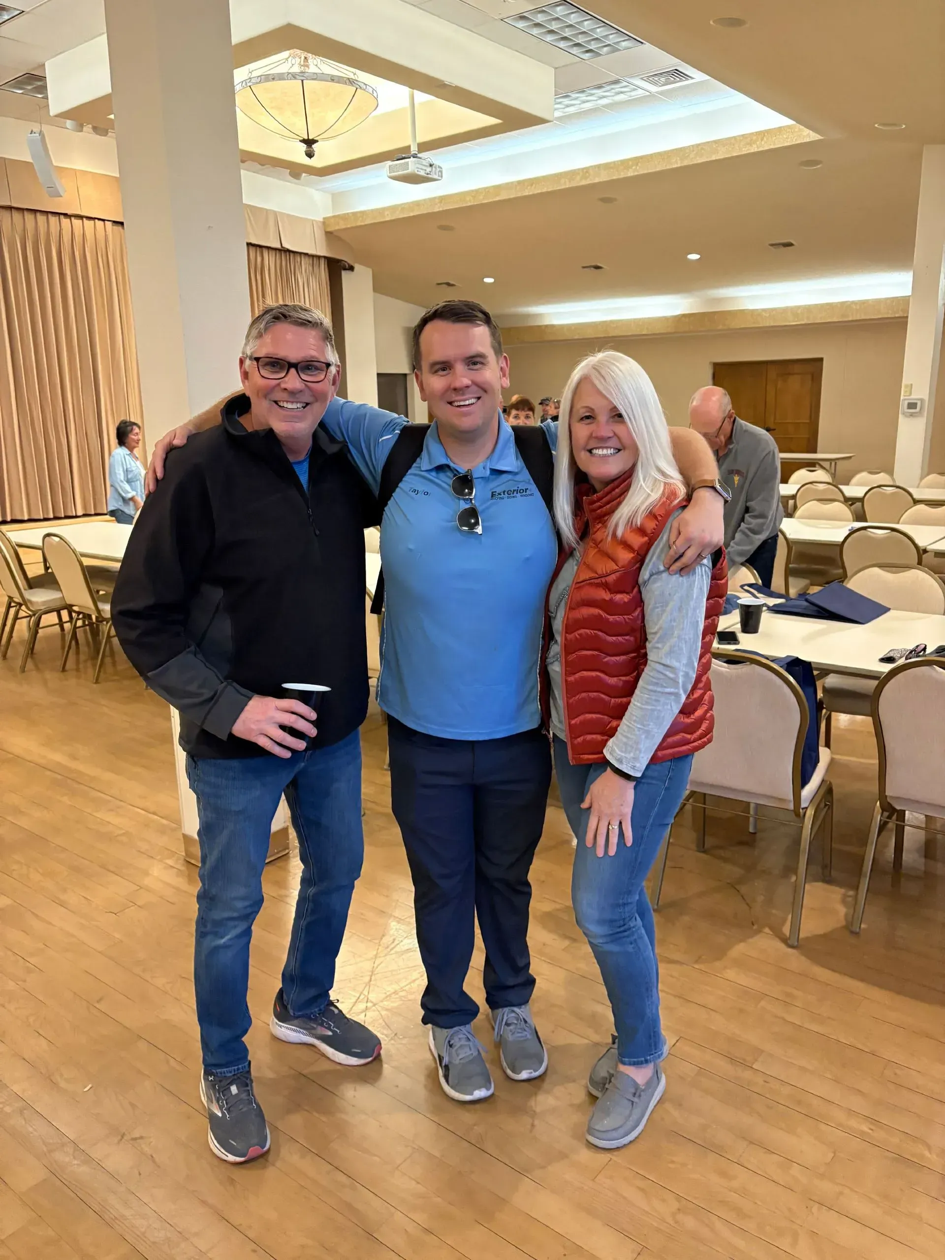 Three people posing together in a bright banquet hall, smiling with arms around each other.