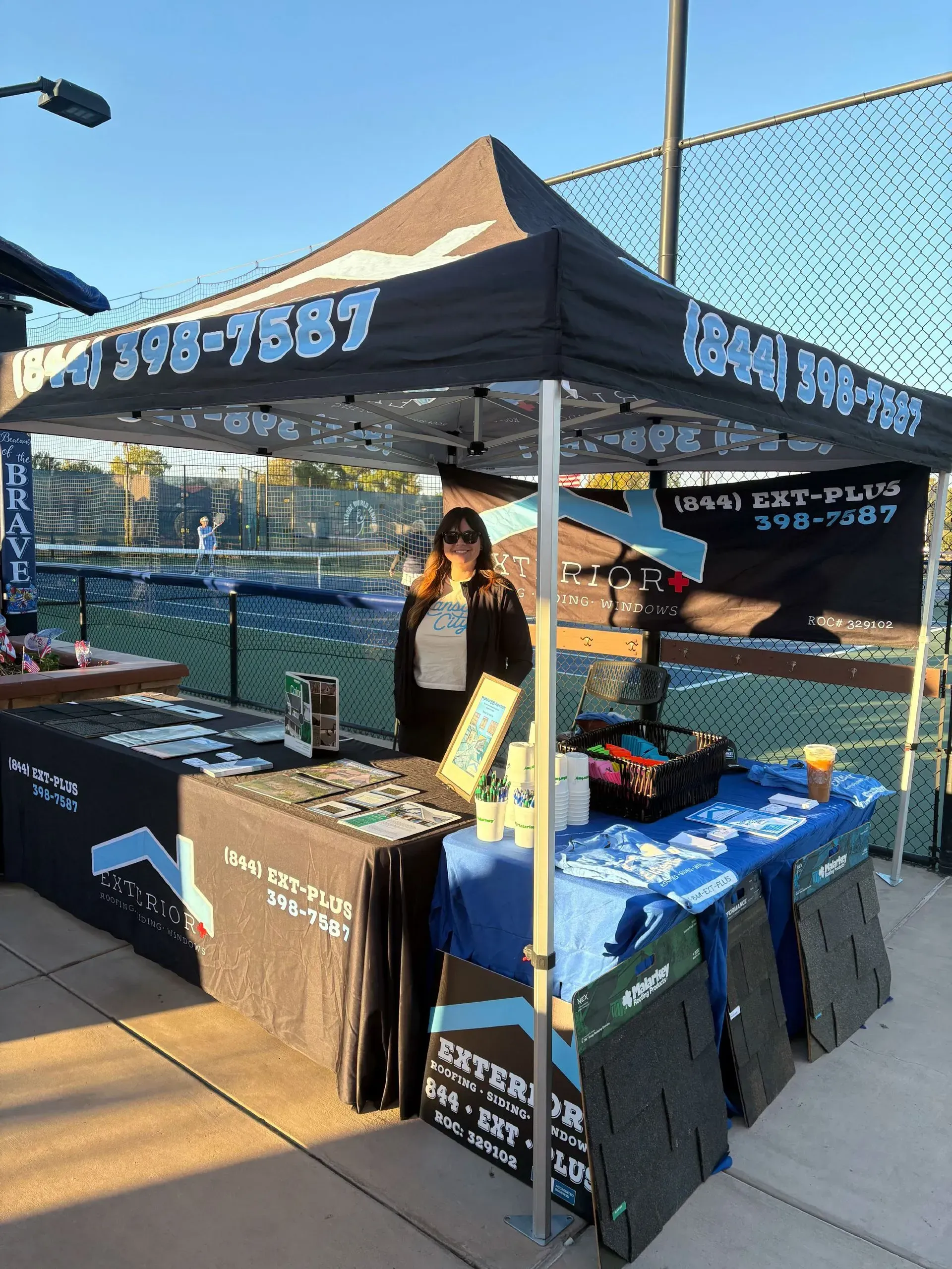 Outdoor vendor booth with black canopy and blue tablecloths, displaying items beside a tennis court.