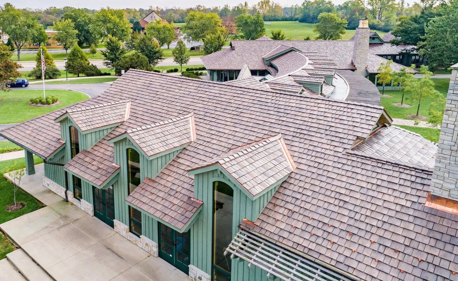 Aerial view of a shingled roof with dormers and a chimney, surrounded by green trees and lawn