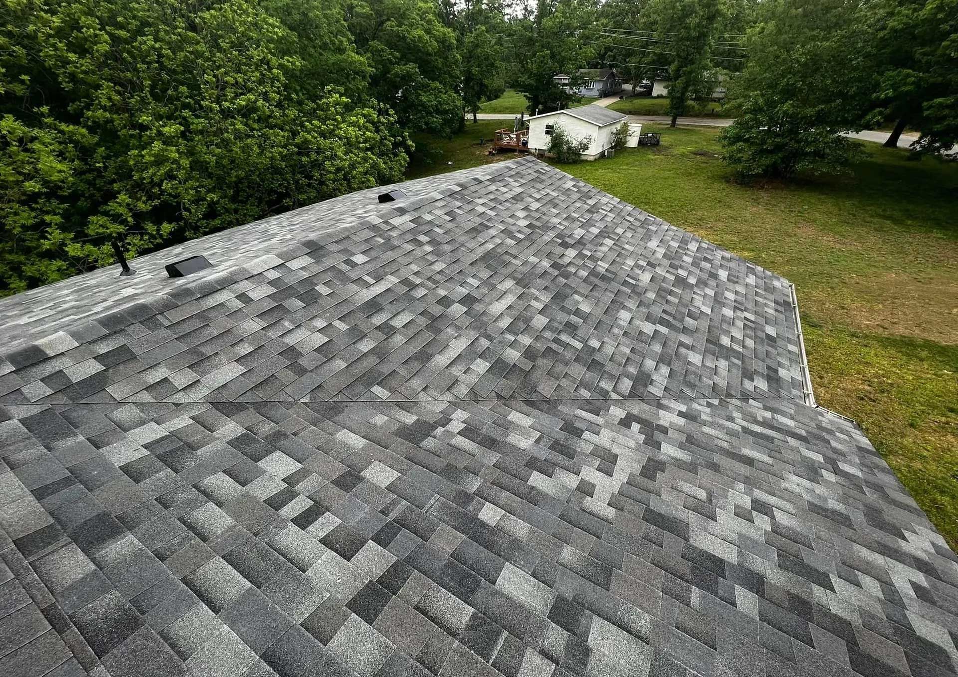 Gray shingled roof in foreground with green trees and yard in the background
