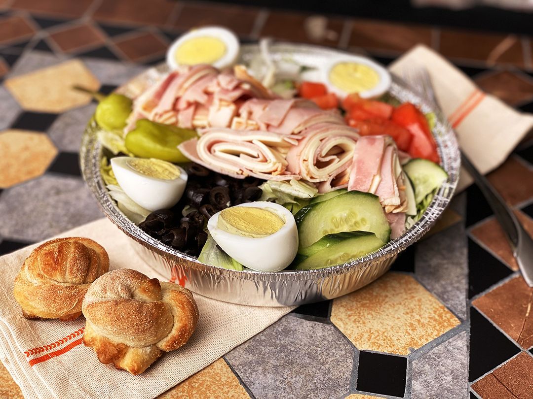 A salad with ham, eggs, cucumbers and olives in a tin foil container on a table.
