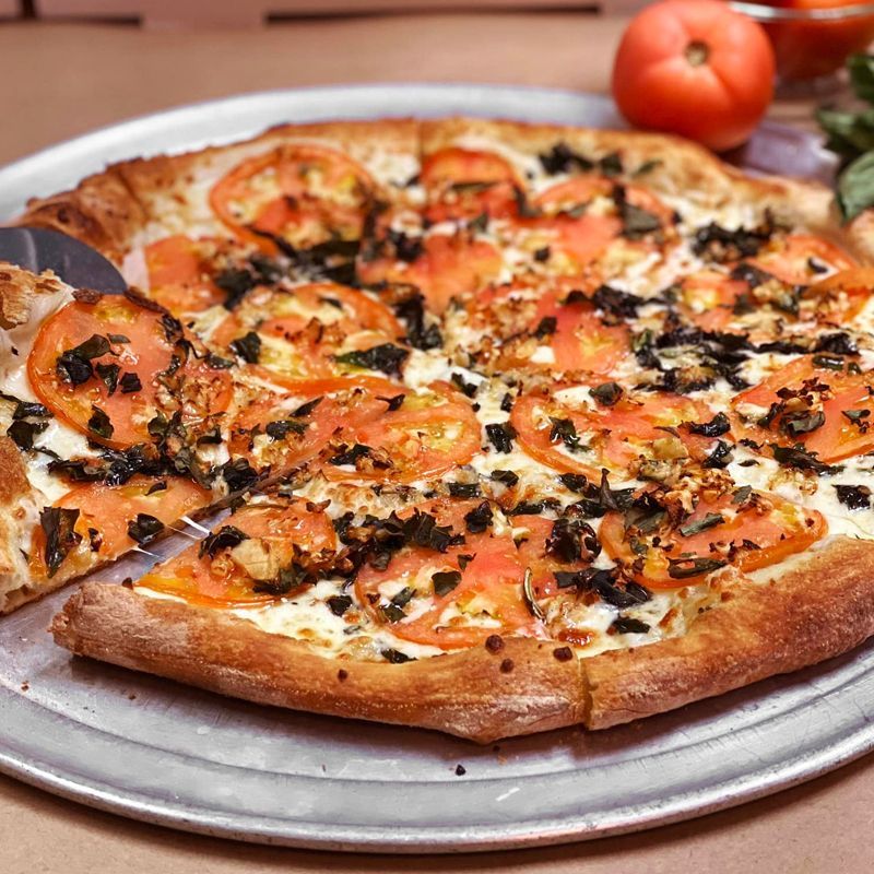 A pizza with tomatoes and cheese is on a pan with a spatula.