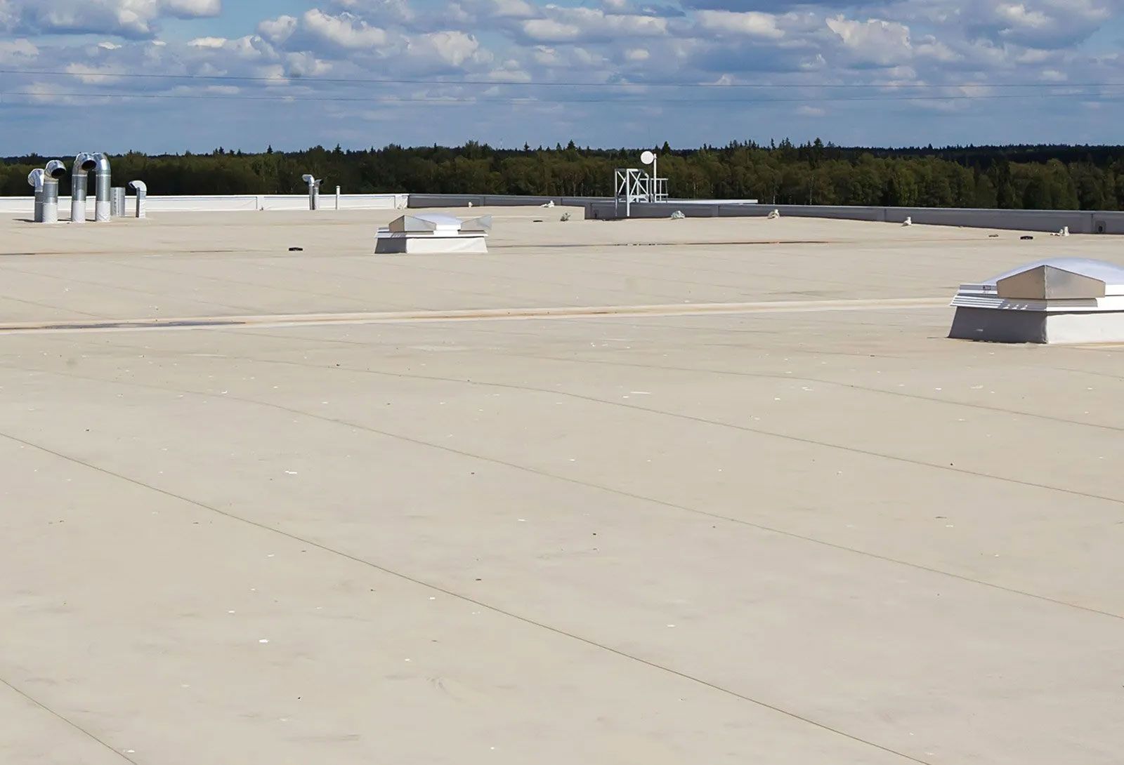 Flat, light-colored commercial roof with vents and skylights. Green trees and blue sky with clouds in the background.